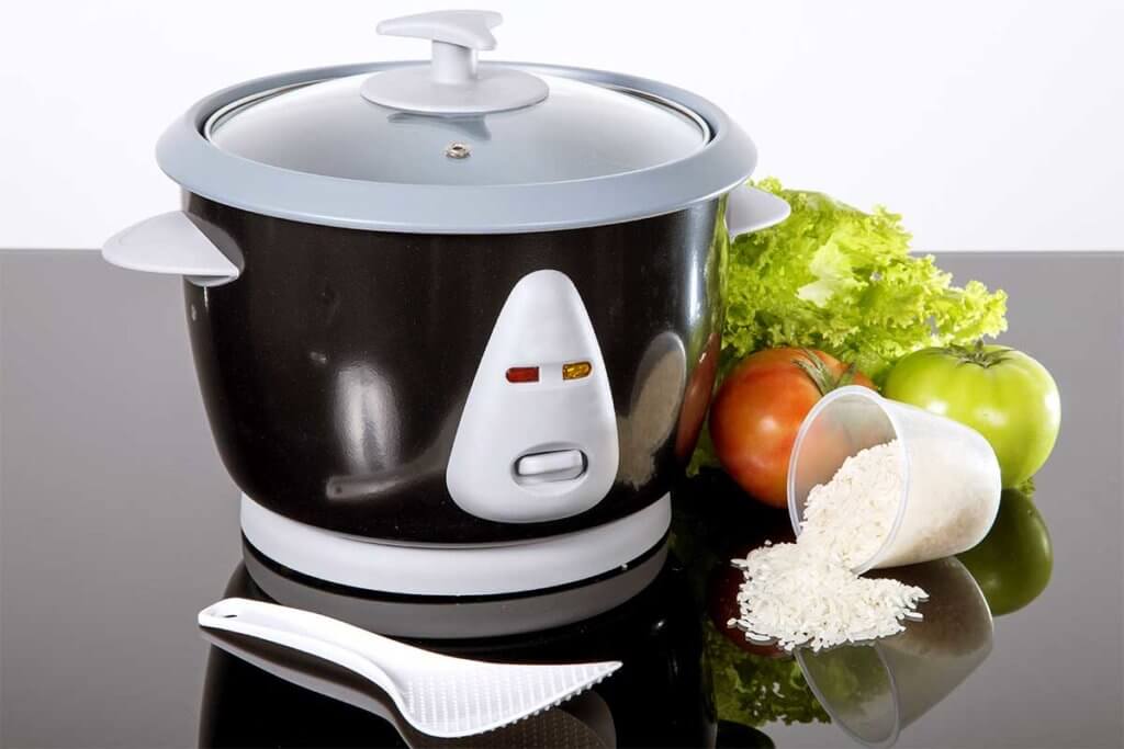 Reiskocher steht auf schwarzem Untergrund, daneben sind Reis und Tomaten platziert